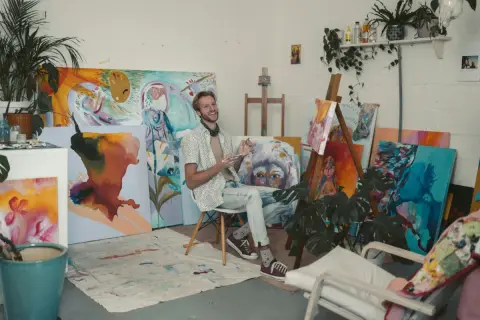 A smiling artist sits in his vibrant artist studio, surrounded by colorful canvases and plants.