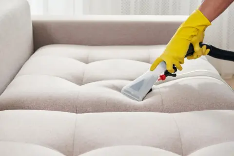 Gelbe Handschuhe führen ein Reinigungsgerät über ein helles Sofa. Tiefenreinigung für ein makelloses Sofa.