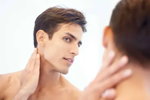 Un homme au visage fin et à la mâchoire marquée, la main caressant son cou, révélant une belle v line.
