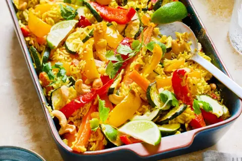 Reisgericht mit buntem Gemüse, Cashewkernen und Limettenspalten in einer Auflaufform.