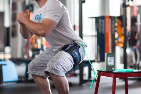 Un athlète en plein squat, équipé d'un appareil de bfr kiné à la cuisse, mesure ses performances.