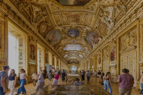 Prunkvolle Deckenmalereien und goldene Verzierungen im Louvre von innen. Besucher bestaunen die Kunstwerke in der langen Galerie.