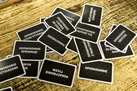 Karty do gry "Czółko" online leżą na drewnianym stole. Zadania na kartach to m.in. "Opowiadanie śpiewając", "Pokazywanie z dłonią na głowie", "Naśladowanie dźwięków".