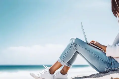 Junge Frau mit Laptop am Strand, genießt die Sonne und das Meer. Ein perfekter Ort für ihr IU Erasmus-Projekt.