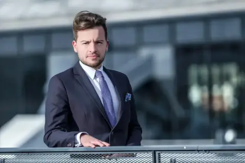 Mężczyzna w garniturze i krawacie opiera się o barierkę na tle nowoczesnego budynku.