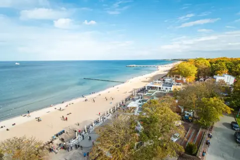Słoneczny dzień na kołobrzeskim deptaku. Ludzie spacerują po plaży i promenadzie, ciesząc się widokiem morza i jesiennych drzew.