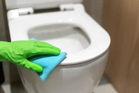 Rękawica w zielonej gumie czyści deskę sedesową niebieską ściereczką. Dowiedz się, czym wyczyścić deskę sedesową, aby była higienicznie czysta.