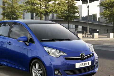 Niebieski Toyota Verso S z czarnym dachem zaparkowany przed nowoczesnym budynkiem.