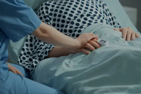 Pielęgniarka trzyma dłoń pacjenta w stanie minimalnej świadomości, rokowania niepewne. Pulsoksymetr na palcu.