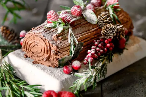 Ein köstliches Bûche de Noël Rezept Original: Schokoladenrolle mit Beeren, Tannenzapfen und Rosmarin, bestäubt mit Puderzucker.