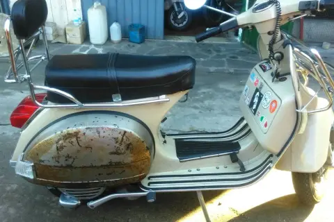Vespa color crema con evidente effetto ruggine sul parafango posteriore. La sella è nera, con portapacchi cromato.