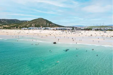 Playa de Zahara de los Atunes con gente bañándose y disfrutando del sol, con el pueblo blanco al fondo y molinos de viento en la colina.