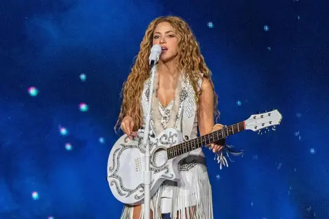 Shakira śpiewa i gra na gitarze podczas koncertu w Polsce. Wokalistka ma długie, falowane włosy i biały, zdobiony strój.