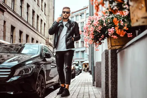 Mężczyzna w czarnej skórzanej kurtce i zniszczonych jeansach, rozmawiający przez telefon, obok lśniącego czarnego samochodu. Idealne buty do kurtki skórzanej dopełniają stylizację.