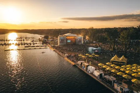 Zachód słońca nad jeziorem, gdzie odbywa się festiwal. Tłumy ludzi bawią się przy scenie, a nad wodą cumują łodzie. To miejsce, gdzie rodzi się sława rap stacja.