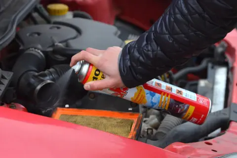 Ręka trzyma puszkę z preparatem do rozruchu silnika, rozpylając go w okolicy filtra powietrza. Czy odpalanie na plaka jest szkodliwe?