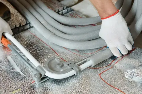 Ręce w białych rękawiczkach zaginają rury PEX za pomocą giętarki, przygotowując je do łączenia w posadzce.