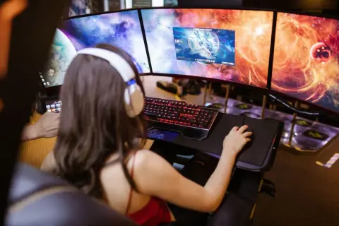 Frau mit Kopfhörern spielt an drei Monitoren. Ein "pile of shame" an Aufgaben wartet, doch jetzt zählt nur das Spiel.