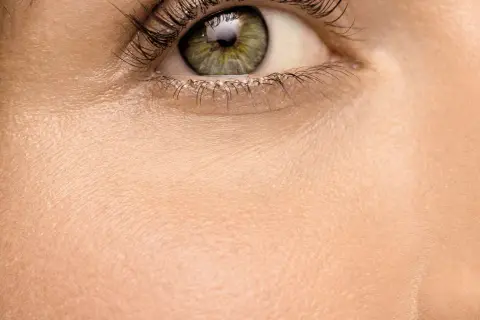 Nahaufnahme eines Auges mit grüner Iris und langen Wimpern. Die Haut wirkt blass, fast wie eine fahle Gesichtsfarbe, und zeigt feine Poren.