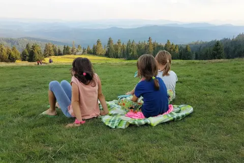 Trzy dziewczynki siedzą na kocu na trawie, podziwiając rozległy widok na Beskid Żywiecki.