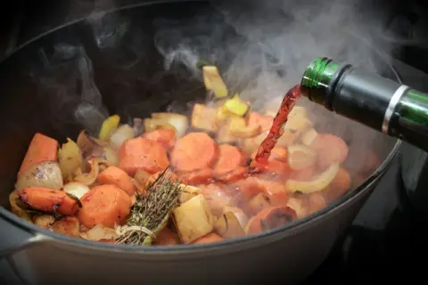 Verduras cocinándose en una olla, se vierte vino tinto. El vapor sube, indicando que el alcohol se evapora rápido.