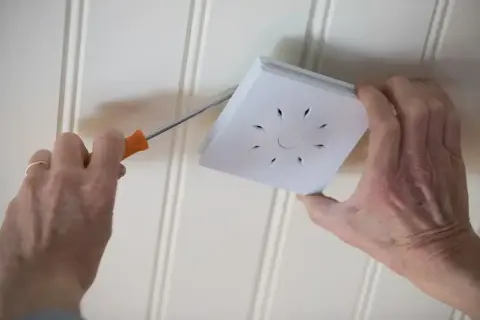 Main tenant répare un détecteur de fumée qui se déclenche sans raison. Il utilise un tournevis pour le démonter.