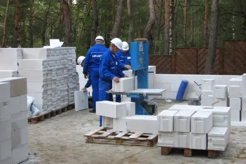 Budowlańcy pracują przy maszynie do cięcia bloczków betonowych. W tle widać stosy materiału budowlanego, co pozwala oszacować, ile bloczków betonowych wchodzi na metr kwadratowy.