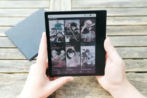 Mains tenant une liseuse affichant des mises à jour de mangas, dont Spy x Family, Kagurabachi et One Piece, sur un fond de table en bois. Le kaleido 3 offre une lecture claire.