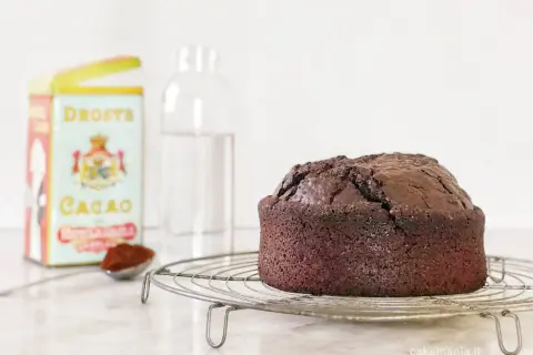 Torta all'acqua al cacao appena sfornata, su una gratella. Accanto, una scatola di cacao Droste e un bicchiere d'acqua.