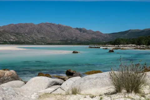 Aguas turquesas y arena blanca bordean la costa, con el imponente monte Pindo como telón de fondo. Rocas cubiertas de musgo salpican la orilla.