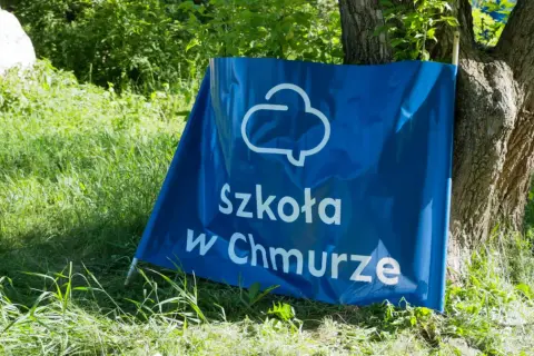 Niebieski baner z napisem "Szkoła w Chmurze" oparty o drzewo, na tle zielonej trawy i liści.