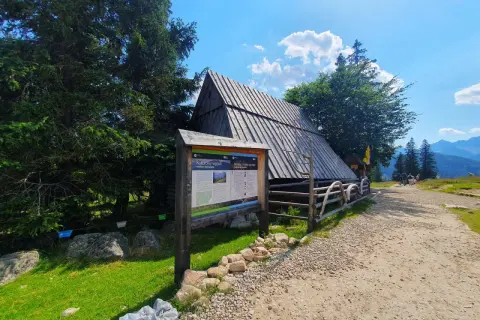 Drewniana chata na górskim szlaku, obok tablica informacyjna o kulturowym wypasie owiec. W oddali widać wierch Poroniec.
