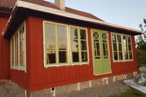 Ett rött hus med pågående arbete för att bygga glasveranda. Gröna fönster och dörrar reflekterar himlen.