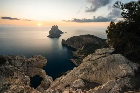 Vista del ojo de es Vedrà al atardecer, con el sol poniente tiñendo el cielo de naranja y el mar en calma.