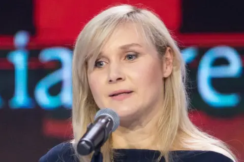 Kobieta z blond włosami mówi do mikrofonu na tle ekranu z napisem.