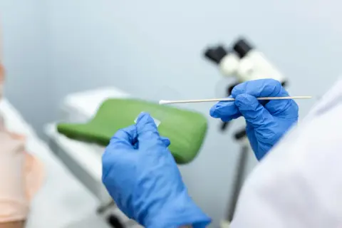 Ręce w niebieskich rękawiczkach przygotowują próbkę do badań cytologia wyniki. W tle mikroskop i fotel ginekologiczny.