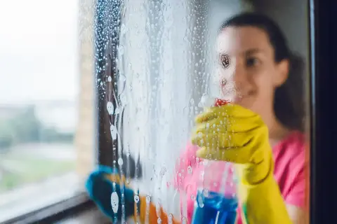 Frau mit gelben Handschuhen sprüht Reinigungsmittel auf ein Fenster, um es zu putzen.
