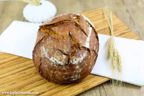 Ein rustikales Bierbrot mit Sauerteig, auf einem Holzbrett mit einem weißen Tuch und Ähren dekoriert.
