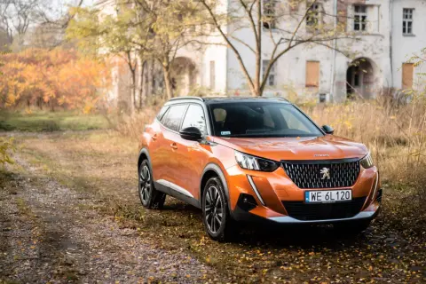 Pomarańczowy Peugeot 2008 GT na tle jesiennego krajobrazu z opuszczonym budynkiem.