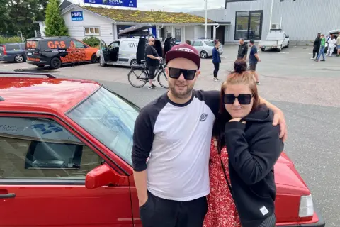 Ett par poserar framför en röd bil, redo att bygga ljud i bil. I bakgrunden syns bilar och folk vid en butik.