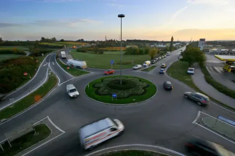 Ruchliwe rondo z wieloma rodzajami rond, samochodami i ciężarówkami. W tle widać pola i niebo o zachodzie słońca.