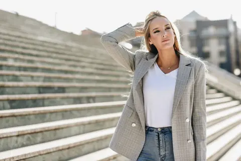 Chiara Ferragni w szarej marynarce w kratę i jeansach, z ręką we włosach, na tle schodów.