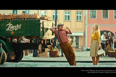Mężczyzna w czapce i szelkach opiera się o ciężarówkę z piwem Žatecký. Ile procent ma to piwo? Kobieta w żółtej bluzce patrzy w górę.