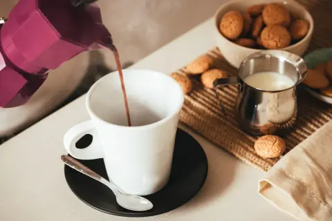 Un buon caffè fumante viene versato da una moka viola in una tazza bianca, accompagnato da biscotti e latte.