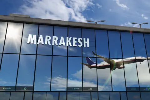 Samolot wznosi się na tle błękitnego nieba z chmurami, odbijając się w szklanej fasadzie lotniska w Marrakeszu.