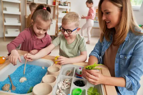 Children explore blue sand and shells in a sensory table, guided by a smiling teacher.