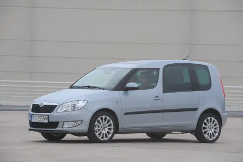 Jasnoszary Skoda Roomster z kierowcą za kierownicą stoi na betonowym placu przed szarym budynkiem.