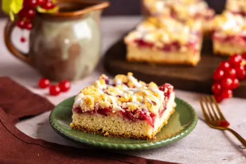 Saftiger Johannisbeerkuchen, wie ihn Landfrauen backen, mit Streuseln und Zuckerguss. Ein Stück auf grünem Teller, daneben rote Johannisbeeren.
