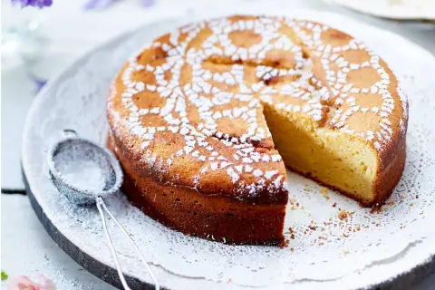 Ein saftiger Joghurt Kuchen, mit Puderzucker bestäubt und einem Stück herausgeschnitten. Daneben liegt ein kleines Sieb.