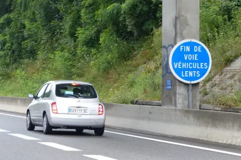 Voiture argentée sur l'autoroute, passant devant un panneau "Fin de voie véhicules lents". La nature luxuriante borde la route.